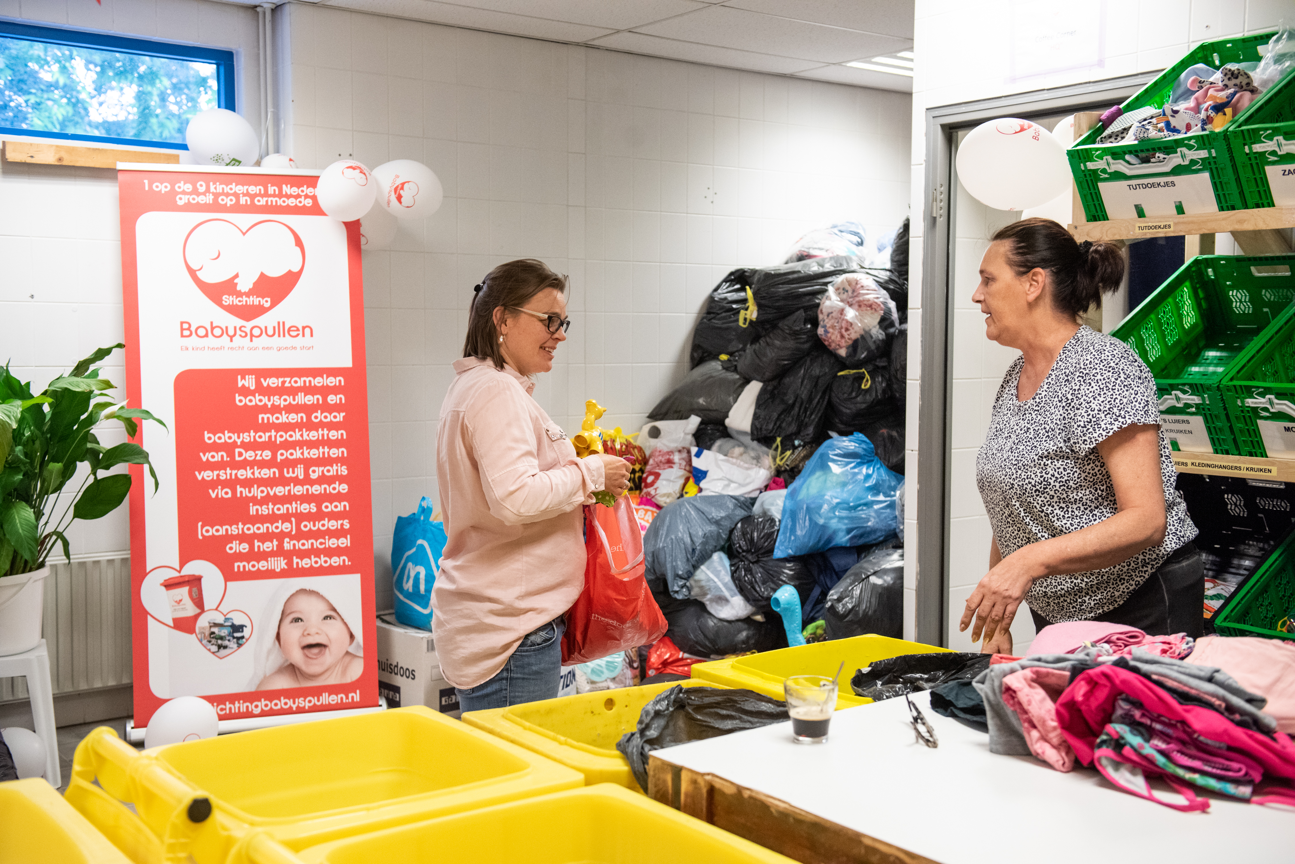 twee vrijwilligers staan voor de sorteerbakken met op de achtergrond een hoge stapel vuilniszakken met gedoneerde babyspullen